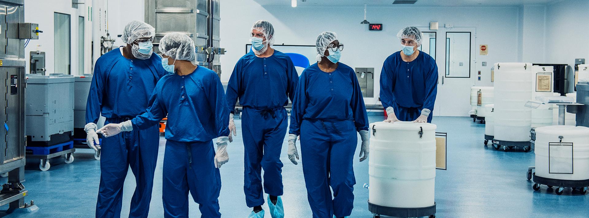   Pfizer Workers in blue sterile suits collaborate in a cleanroom with large white storage containers.
