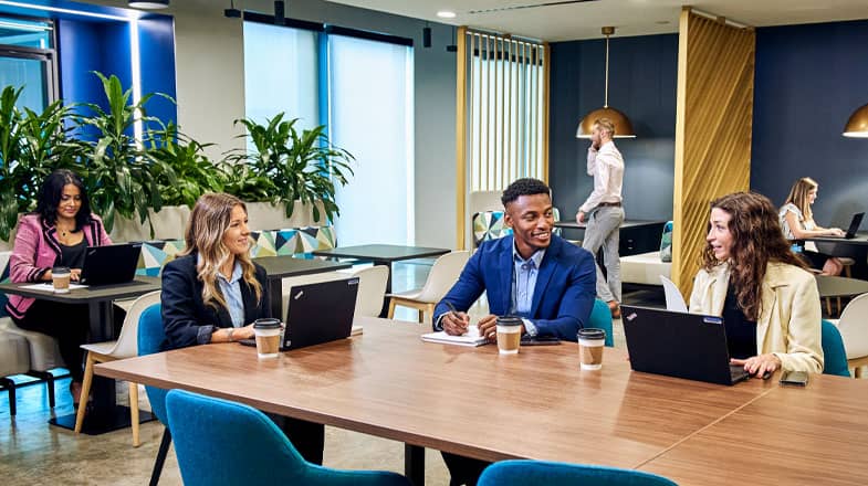 Photograph of coworkers at laptops talking around a large shared table in a modern office