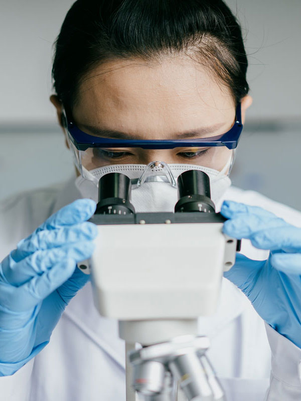 Photograph of a lab technician using a microscope, wearing blue gloves and a lab coat