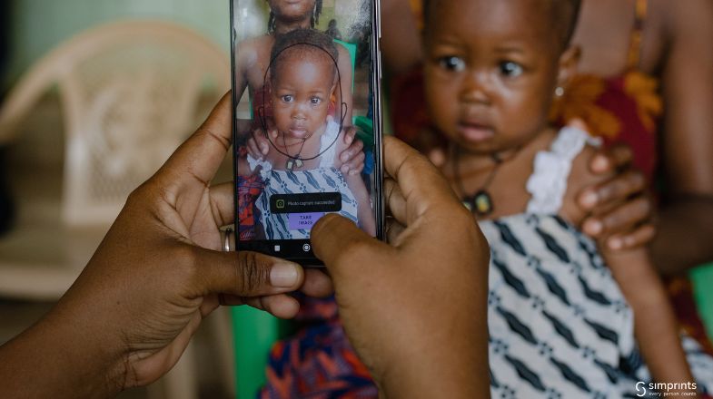 Hands holding a smartphone, photographing a woman in patterned clothing holding a baby