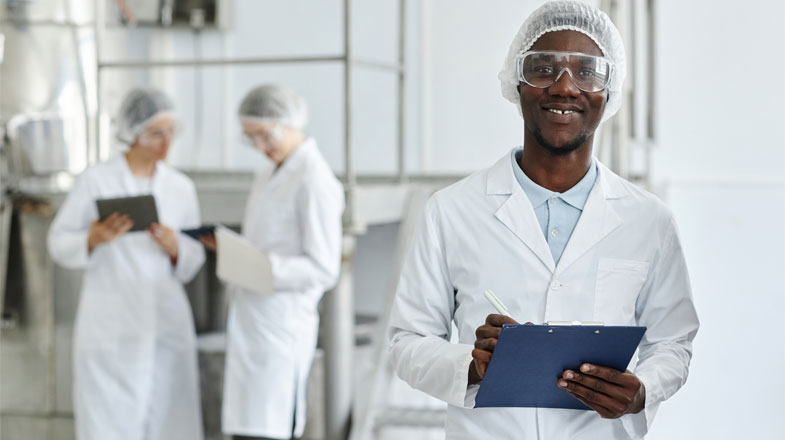Photograph of smiling lab worker with clipboard; two colleagues work in background thumbnail