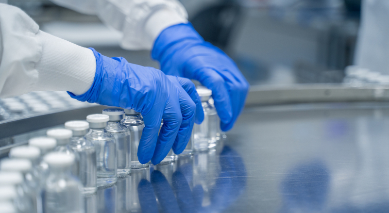 Gloved hands arranging glass vials on a metal conveyor in a laboratory