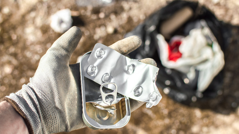 Gloved hand holding a crushed soda can tab near a trash bag outdoors
