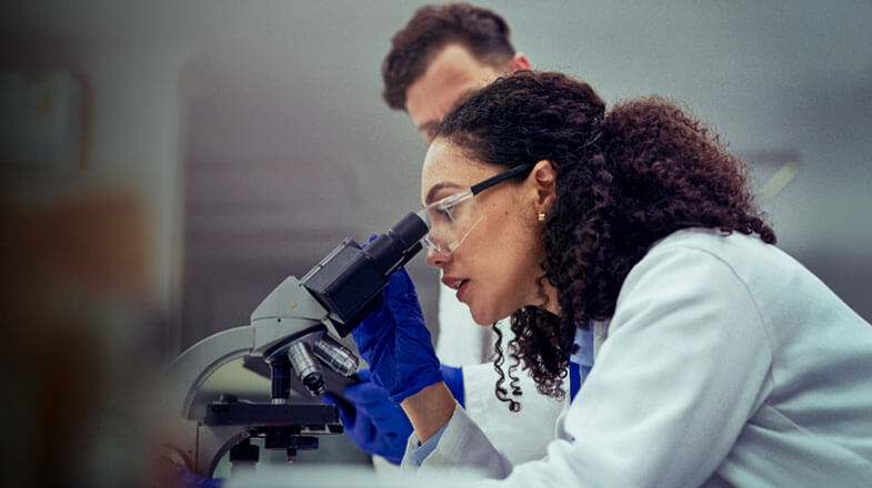 Photograph of a scientist using a microscope, with a colleague standing behind thumbnail