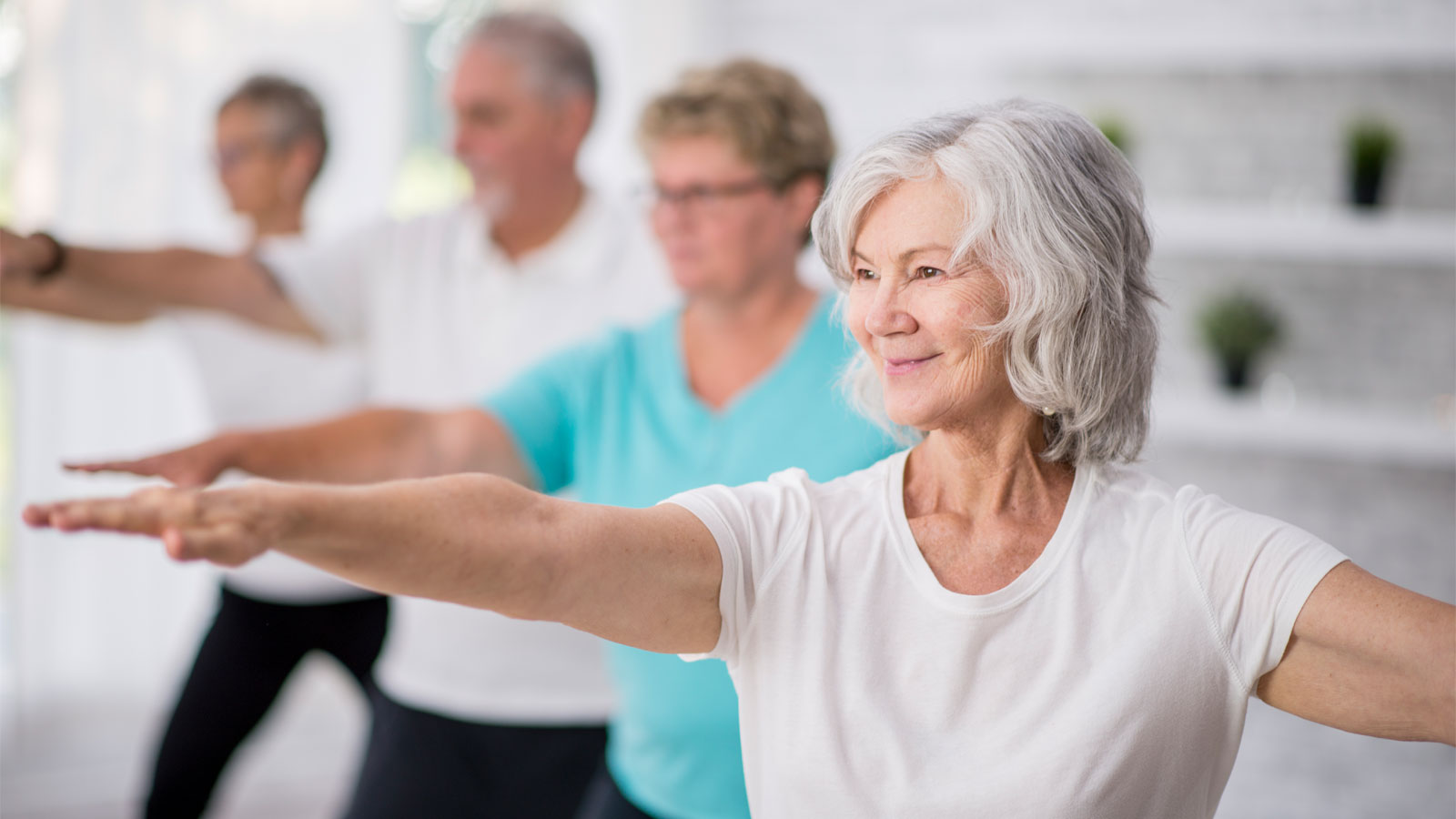 A multi-ethnic group of adult men and women are indoors in a fitness studio. They are wearing casual clothing while at a yoga class. A senior Caucasian woman is smiling while stretching out her arms.