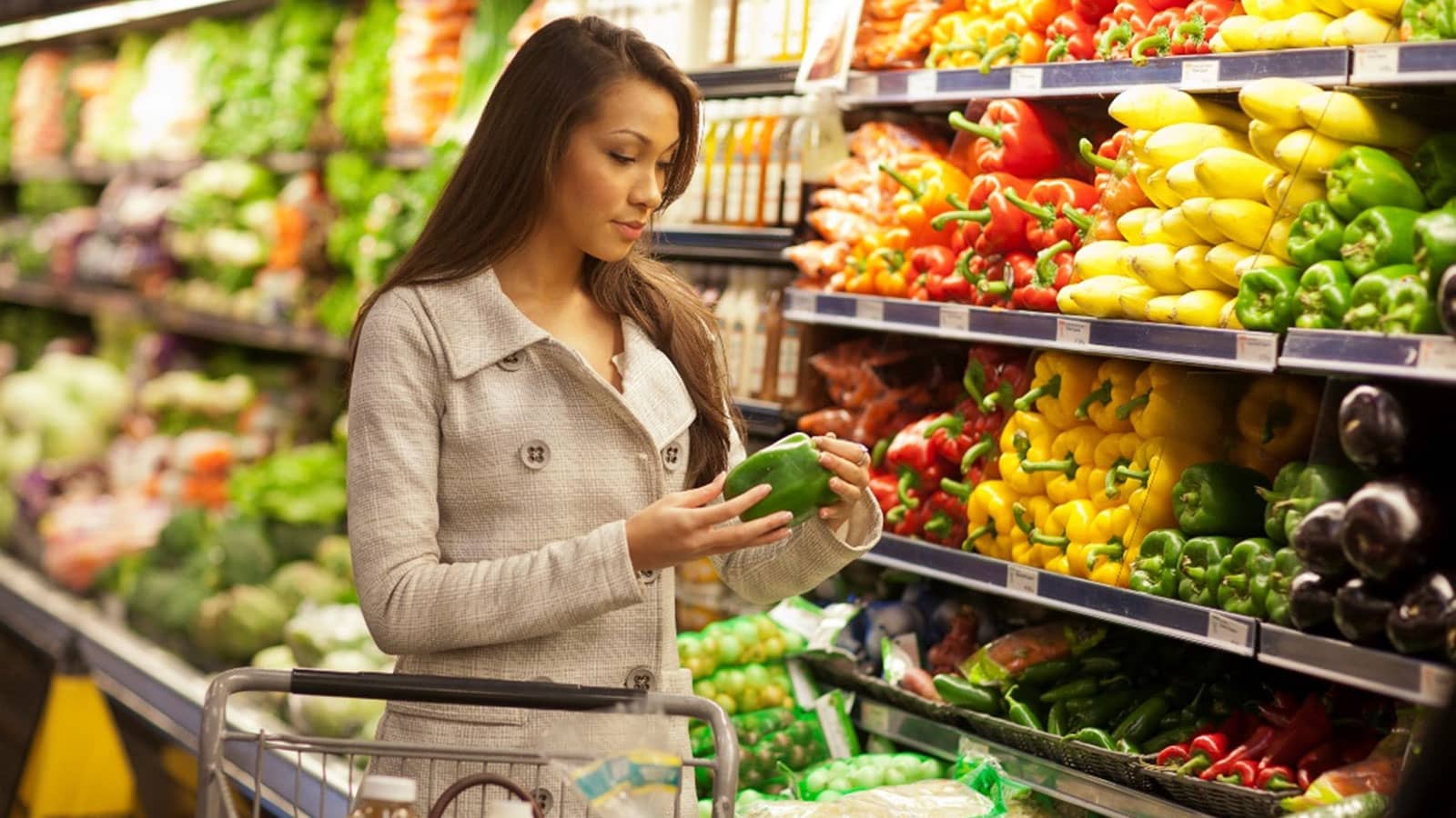 Person examining a green bell pepper in a grocery store produce aisle with colorful vegetables.