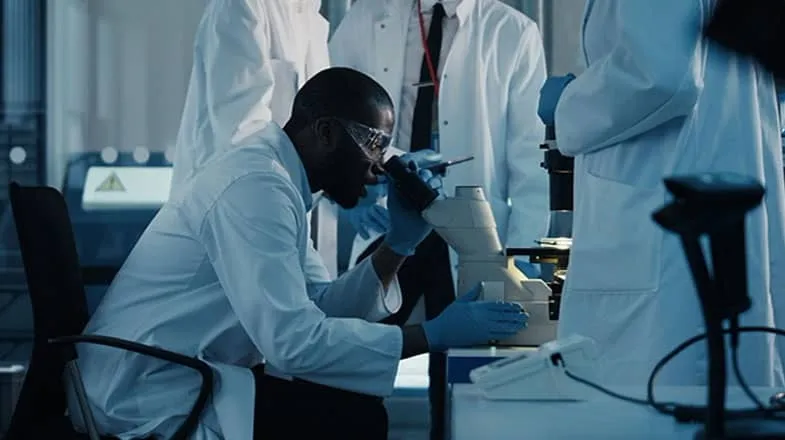 Scientist in lab coat using a microscope, surrounded by colleagues in a laboratory thumbnail