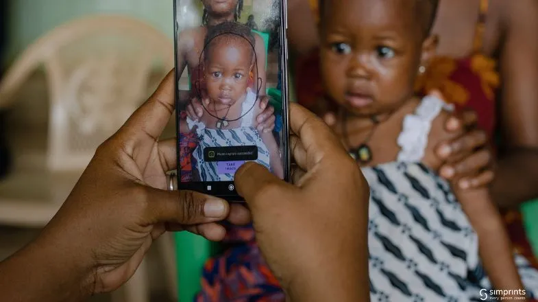 Hands holding a smartphone, photographing a woman in patterned clothing holding a baby