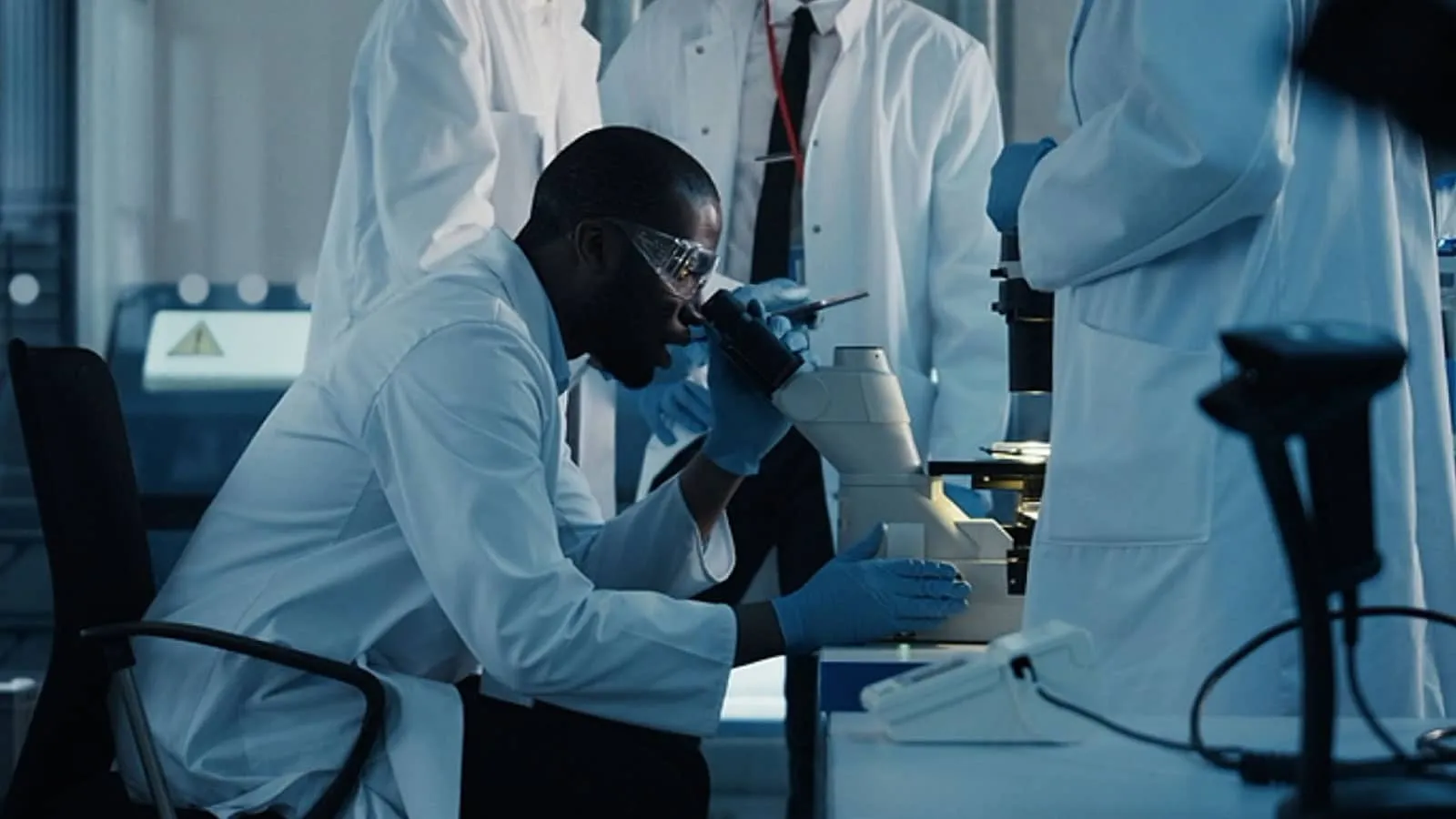 Scientist in lab coat using a microscope, surrounded by colleagues in a laboratory