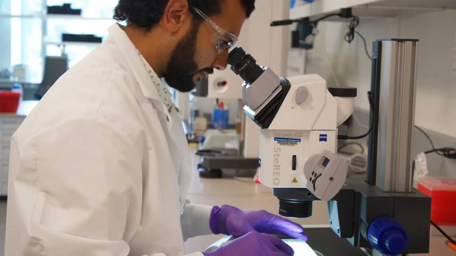 Photograph of a lab worker using a microscope while wearing gloves and a white coat