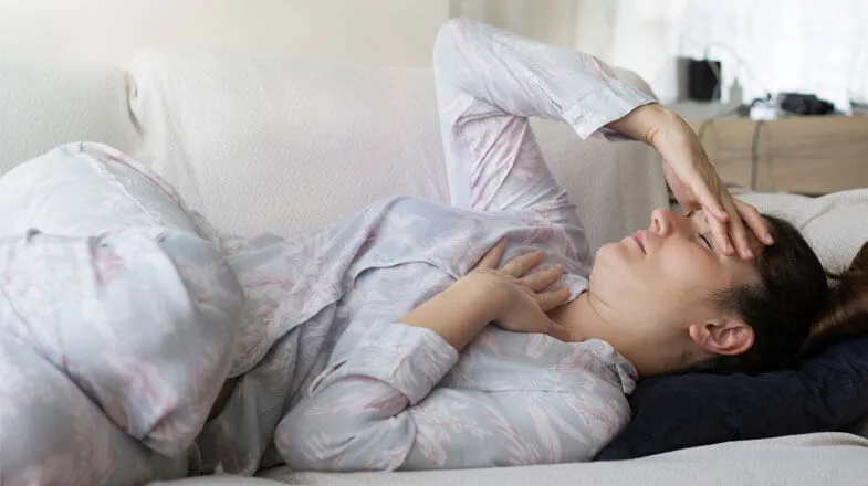 Photograph of a woman in pajamas lying on a couch, holding her head and chest, thumbnail.