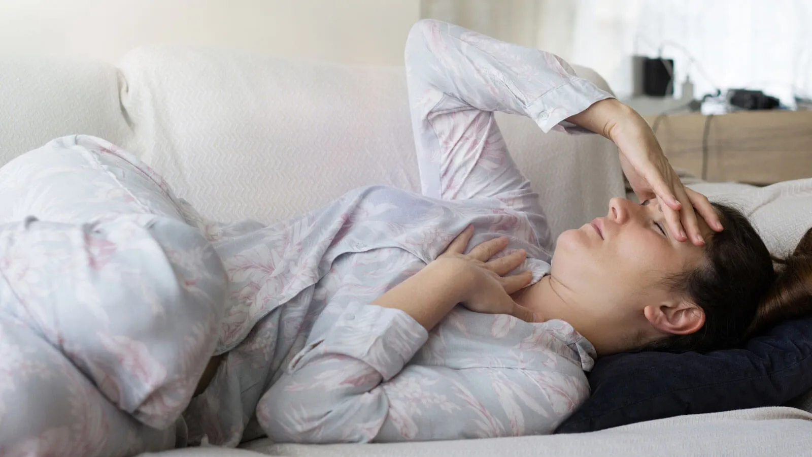 Photograph of a woman in pajamas lying on a couch, holding her head and chest.