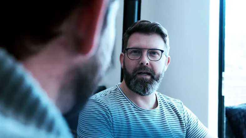 Photograph of a bearded man in glasses talking with another person indoors, thumbnail.