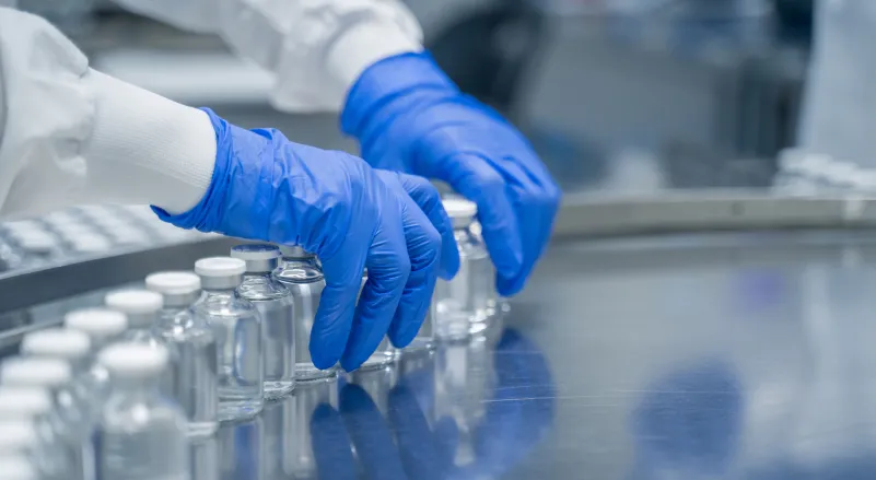 Gloved hands arranging glass vials on a metal conveyor in a laboratory