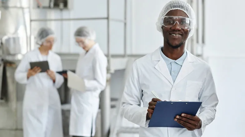 Photograph of smiling lab worker with clipboard; two colleagues work in background