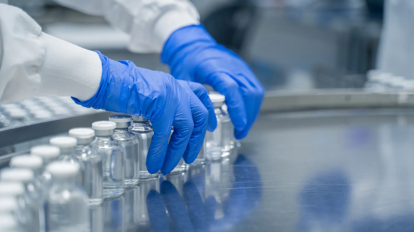 Gloved hands arranging glass vials on a metal conveyor in a laboratory