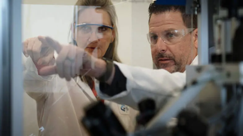Photograph of two scientists in lab coats pointing at equipment behind glass thumbnail