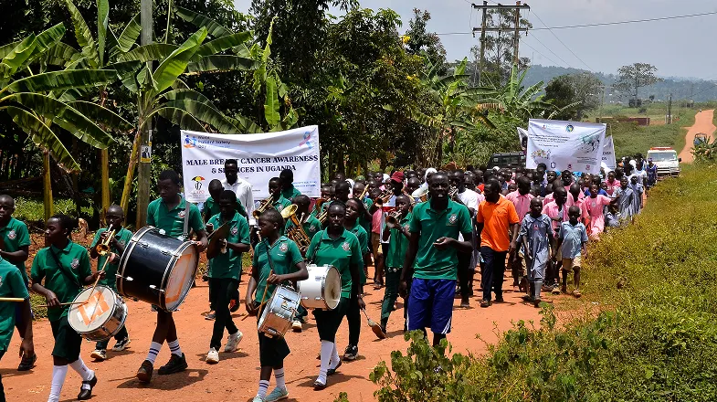 Children and adults marching down a rural road, led by a school band in green uniforms