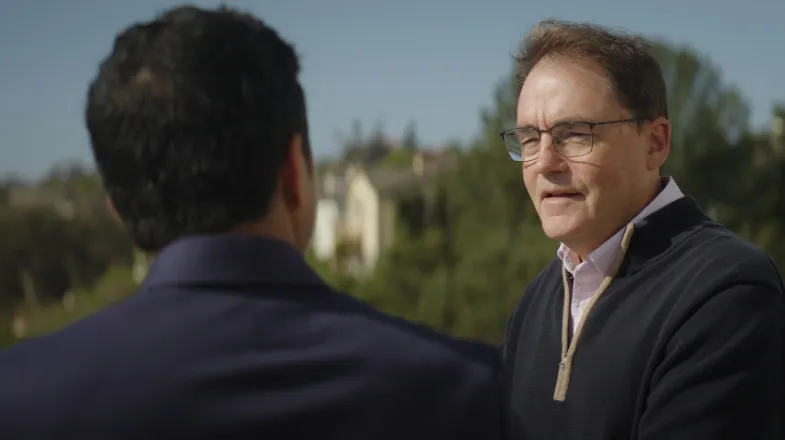 Photograph of two men talking outdoors with trees and buildings in the background