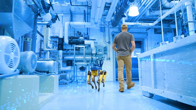 Photograph of a worker and a yellow robot dog walking through a blue-lit industrial plant
