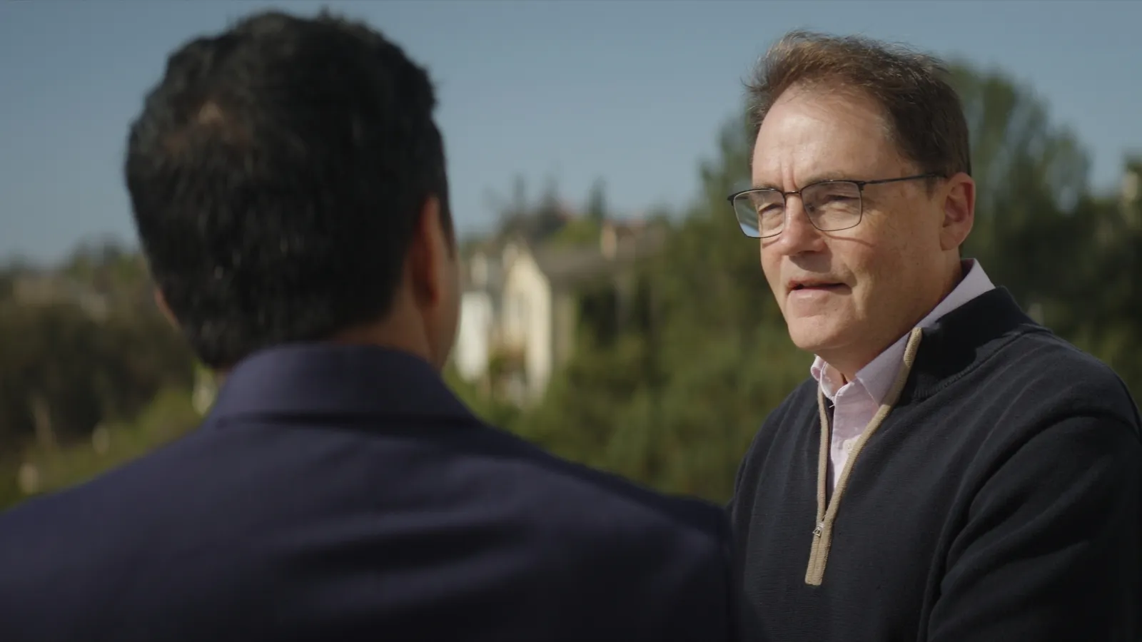 Photograph of two men talking outdoors with trees and buildings in the background