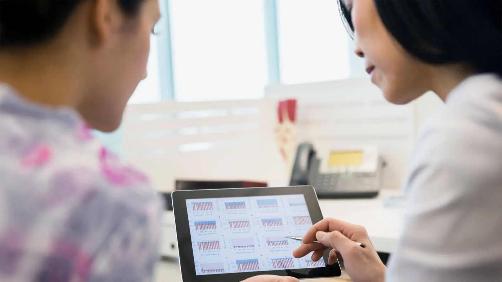 Two women review data charts on a tablet in a bright office setting