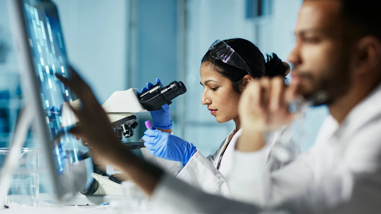 Photograph of lab scientists, one using a microscope, working in a bright laboratory.
