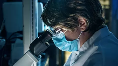 Careers in Science, scientist with PPE mask looking through microscope lens