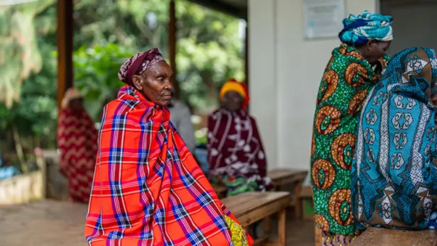Photograph of women in colorful patterned wraps sitting and standing on wooden benches