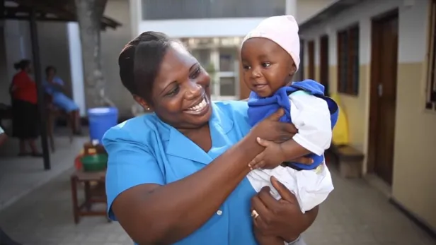 Smiling nurse in blue uniform holding a baby in a courtyard