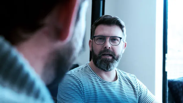 Photograph of a bearded man in glasses talking with another person indoors, thumbnail.