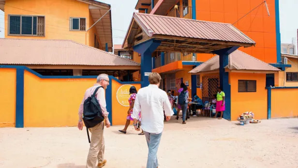 Two men walk toward a bright orange building where people gather near the entrance.