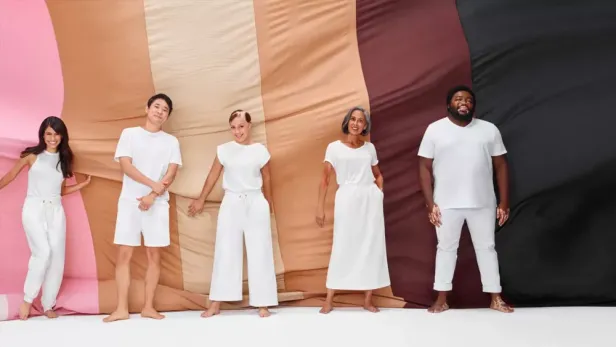 Photograph of five diverse people in white clothes standing before a multicolor skin-tone backdrop