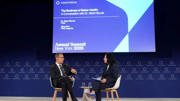 Photograph of two people onstage in chairs talking beneath a large event screen thumbnail
