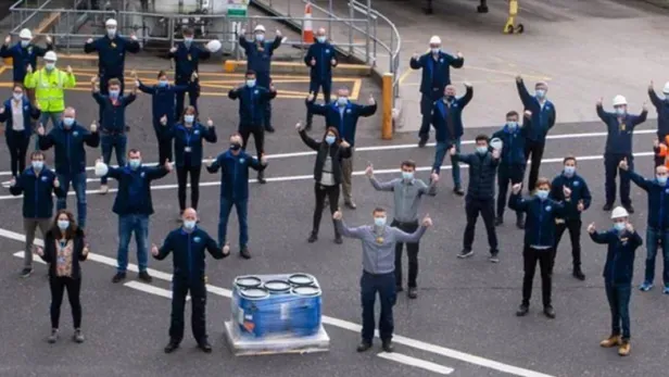 Aerial photo of pfizer masked workers spaced out, raising arms in an industrial yard.