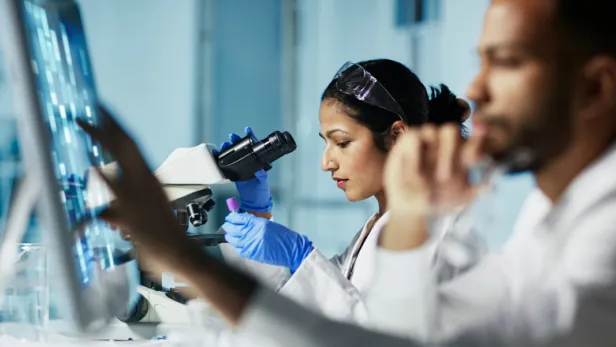 Photograph of lab scientists, one using a microscope, working in a bright laboratory.